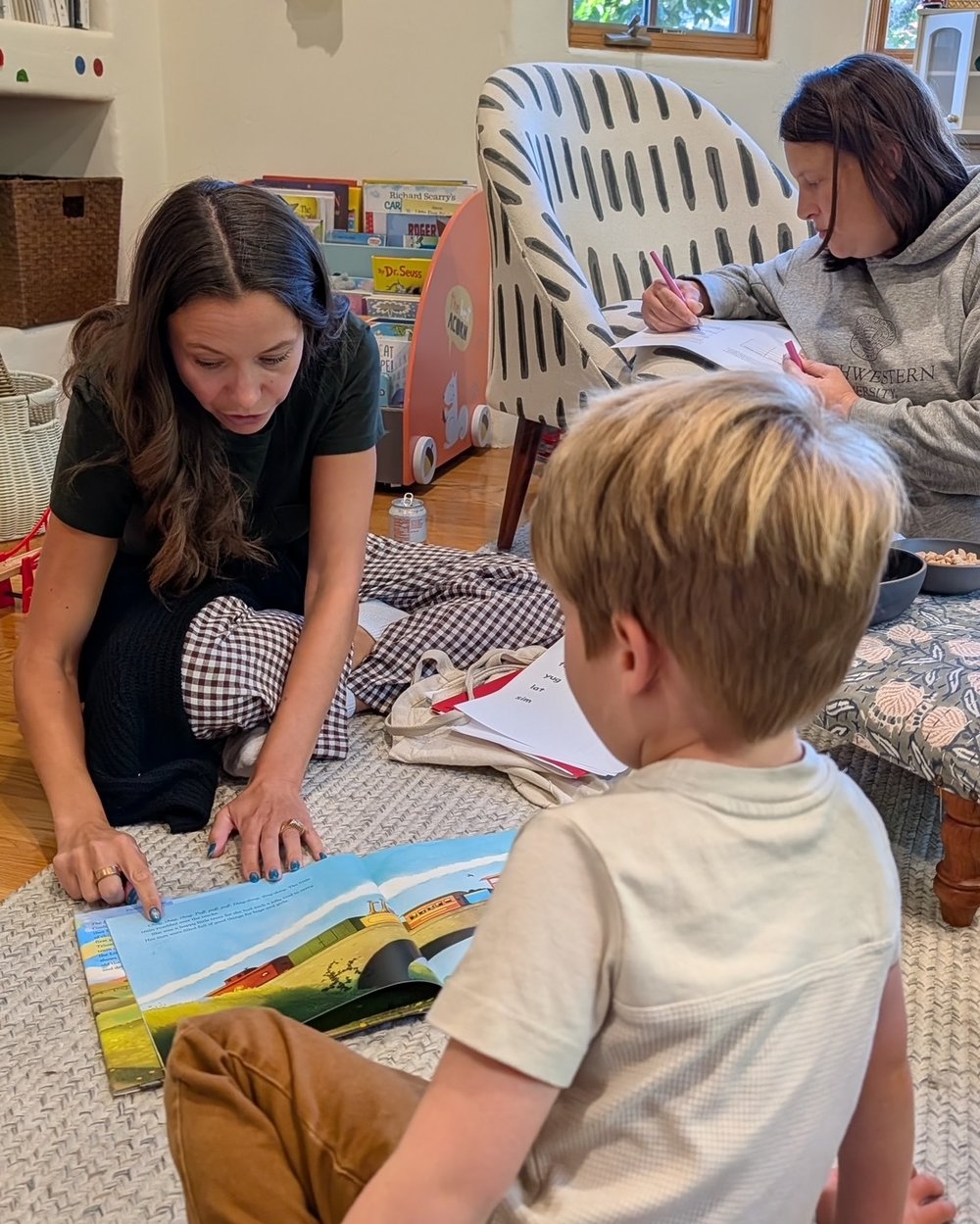 Kaitlyn recording pre assessment notes while a child reads a picture book with his mom on the living room rug